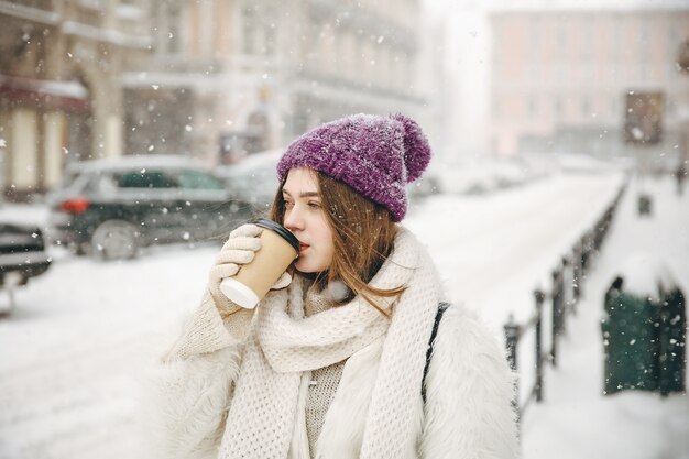 Linda mujer joven bebiendo café recién hecho de taza desechable mientras está de pie al aire libre durante las nevadas.