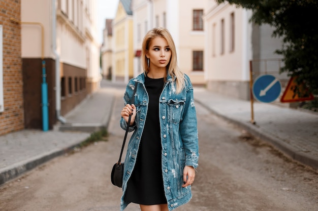 Linda mujer joven y bonita con un elegante vestido negro con un bolso de moda en una chaqueta de mezclilla larga de moda posando en una ciudad en la calle cerca de edificios antiguos