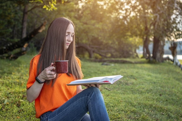 Una linda mujer joven con una camisa naranja está sentada sobre la hierba verde en el bosque con una taza de té y un libro. Concepto de un agradable pasatiempo, descanso, relajación, estilo de vida.