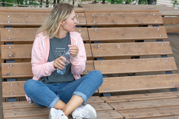 Linda mujer joven caucásica de cuarenta años sentada en una tumbona de madera y bebiendo agua fresca de una botella. Concepto de descanso y relajación.