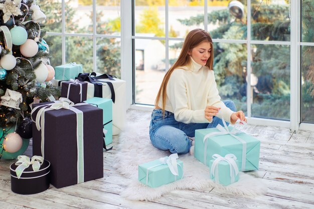 Foto linda mujer joven cerca del árbol de navidad abre los regalos de navidad en una habitación luminosa y acogedora