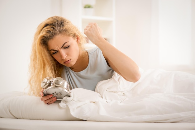 Linda mujer joven despertando por la mañana. Ella está furiosa mirando con despertador.