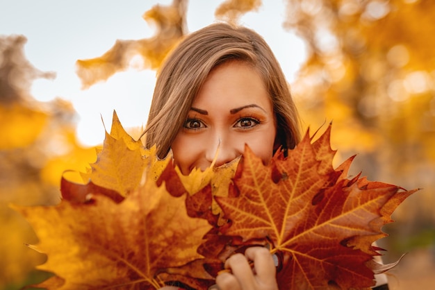 Linda mujer joven disfrutando en el bosque soleado en colores otoñales. Ella sostiene muchas hojas caídas y mira la cámara detrás de ellas.