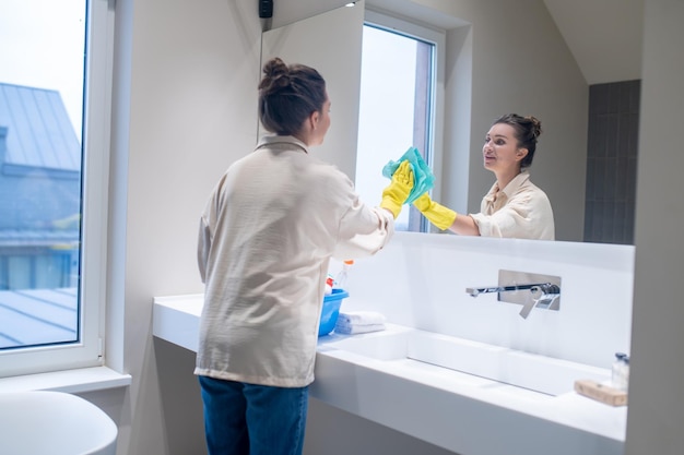 Linda mujer joven limpiando el espejo en el baño