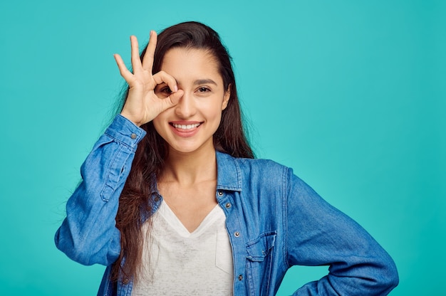 Linda mujer joven muestra ok dign, pared azul, emoción positiva. Expresión facial, persona femenina mirando a cámara en estudio, concepto emocional, sentimientos