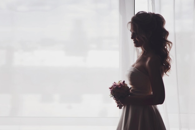 Linda mujer joven novia vestida con ramo de flores en la ventana en la habitación del hotel