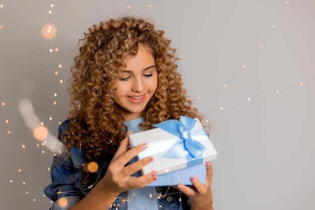 Linda mujer joven con ojos azules y cabello rizado rubio en ropa de mezclilla tiene una caja de regalo en sus manos. chica abre el regalo. fondo gris sólido. espacio para texto. foto de alta calidad