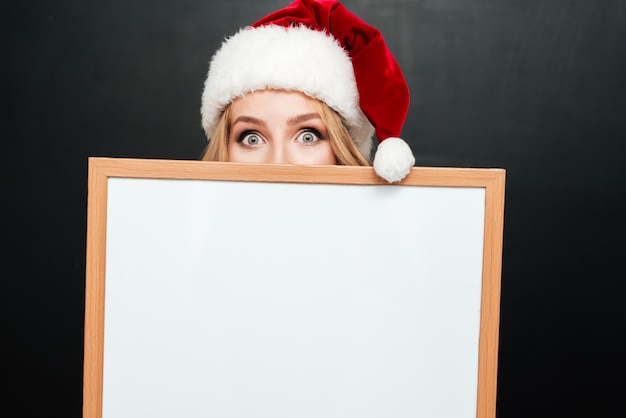 Linda mujer joven con sombrero de santa claus escondiendo la cara detrás de la pizarra en blanco