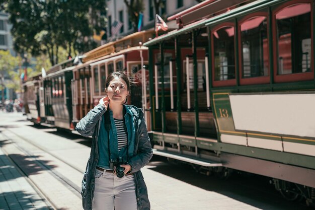 Linda mujer joven sonriente en vacaciones de verano caminando por la calle de la ciudad contra el teleférico en la línea de la calle en san francisco. chica viajera que sostiene una cámara digital se relaja bajo el sol mientras hace turismo urbano