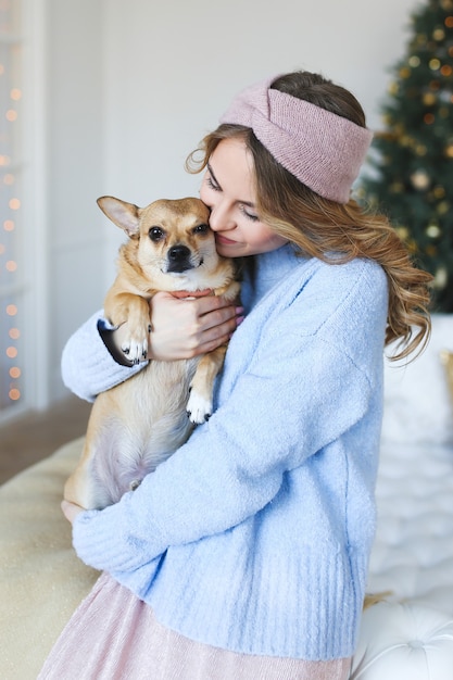 Linda mujer joven en un suéter acogedor con un perro abrazándose y besándose junto al árbol