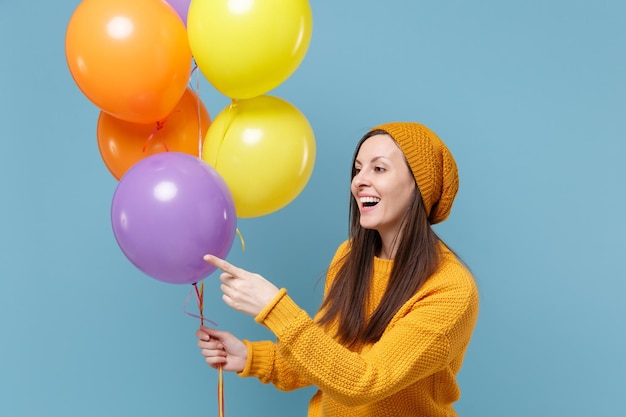 Linda mujer joven en suéter sombrero posando aislado sobre fondo azul. Concepto de emociones de la gente de la fiesta de cumpleaños. Simulacros de espacio de copia. Celebrando sostener globos de aire coloridos señalar con el dedo índice a un lado.