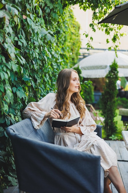 Una linda mujer joven con un vestido se sienta con un cuaderno y hace notas en la terraza de un café de verano Mujer con ropa de verano casual y elegante