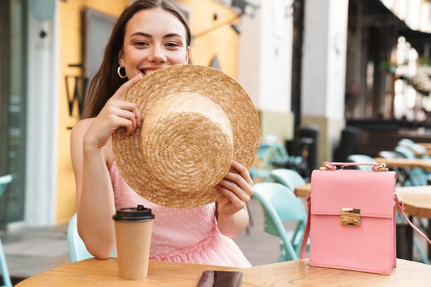 Linda mujer morena sonriendo y sosteniendo un sombrero de paja mientras está sentado en la calle café de verano con café para llevar