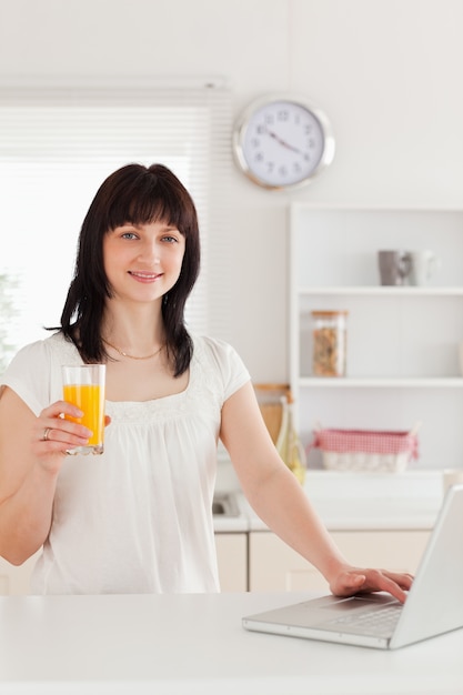 Linda mujer morena con un vaso de jugo de naranja mientras se relaja con su computadora portátil