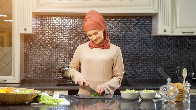 Una linda mujer musulmana con hiyab cocina un delicioso almuerzo para la familia cortando verduras y poniendo condimentos a la luz del sol