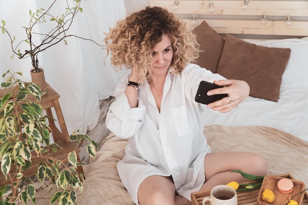 Linda mujer rizada desayunando en la cama en el hotel y haciendo una foto selfie por teléfono