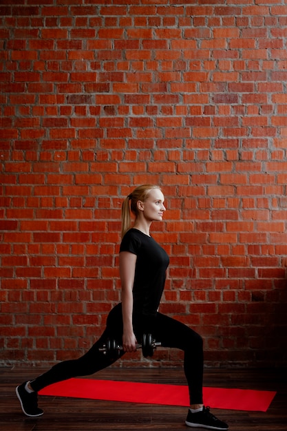 Linda mujer rubia haciendo fitness en el fondo de una pared de ladrillos de la casa durante la cuarentena.