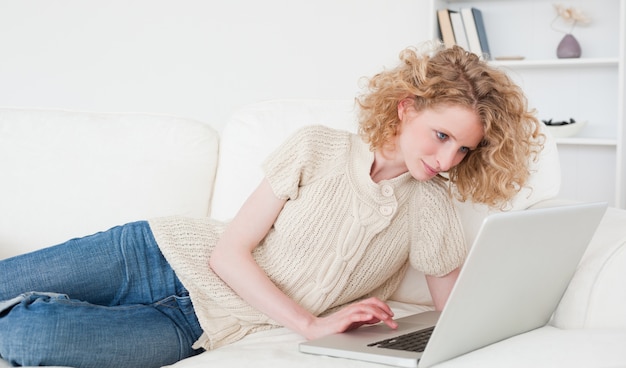Linda mujer rubia relajante con su computadora portátil mientras está acostado en un sofá
