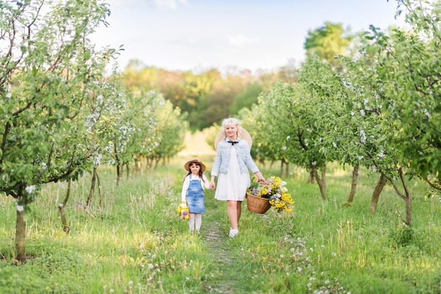 Linda mujer rubia con su pequeña hija en un jardín floreciente de primavera Mamá e hija caminando en la naturaleza con flores en una canasta