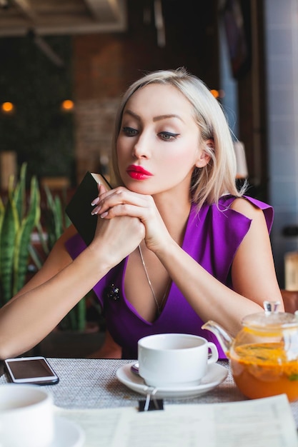 Linda mujer sentada en el restaurante bebiendo té de hierbas soñando y sosteniendo un libro en sus manos