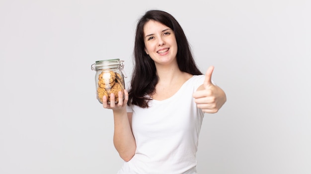 Linda mujer sintiéndose orgullosa sonriendo positivamente con los pulgares hacia arriba y sosteniendo una botella de vidrio de galletas