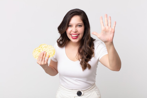 Linda mujer sonriendo y luciendo amigable mostrando el número cinco y sosteniendo una dieta de pasteles de arroz