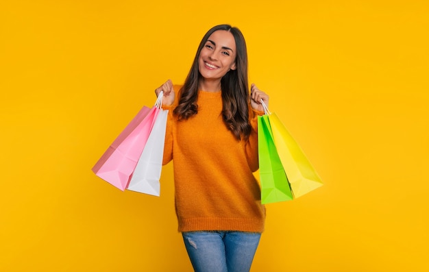 Linda mujer sonriente joven de moda sostiene muchas coloridas bolsas de papel de compras en la mano mientras está aislada sobre fondo amarillo