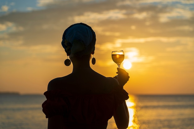 Linda mujer sosteniendo una copa de vino blanco contra una hermosa puesta de sol cerca de las olas del mar en la playa tropical, de cerca. Chica disfruta de la puesta de sol con una copa de vino. Concepto de ocio y viajes