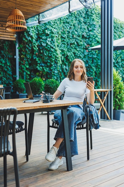 Una linda mujer toma café en la terraza de una cafetería de verano y trabaja en un trabajo remoto con una computadora portátil mientras está de vacaciones en una cafetería con una computadora portátil