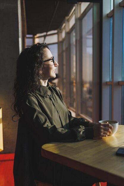 Una linda mujer trabaja en una computadora portátil en un café Joven mujer morena concentrada con gafas sentada en la mesa cerca de la ventana tomando café Trabajo independiente y remoto Estilo de vida moderno de la mujer