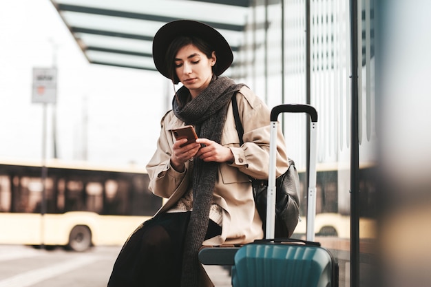 Linda mujer usa un teléfono mientras está sentado en una parada de autobús de la ciudad