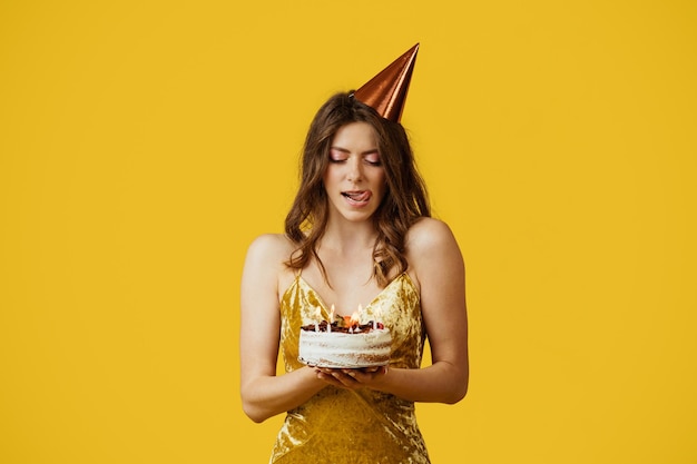 Linda mujer vestida lamiéndose los labios de la tentación mirando un delicioso pastel de cumpleaños con fondo amarillo