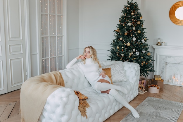 Una linda mujer con un vestido descansa mientras está sentada en un sofá blanco cerca de un árbol de Navidad en un interior claro de una casa acogedora