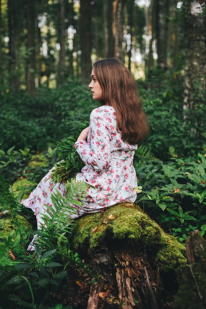 Una linda mujer con un vestido de flores está sentada con un ramo de helechos en el bosque.