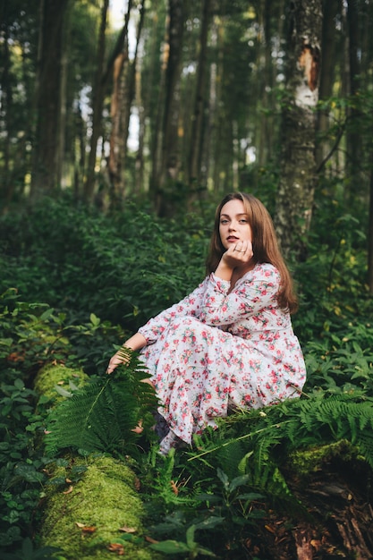 Una linda mujer con un vestido de flores está sentada con un ramo de helechos en el bosque.