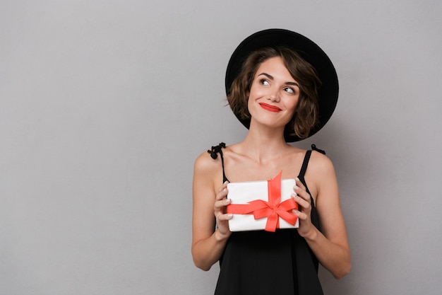 Linda mujer con vestido negro y sombrero con caja de regalo, aislada sobre pared gris