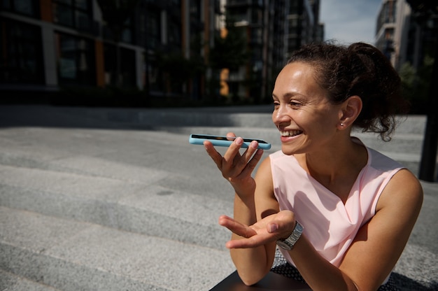 Linda mulher adorável mestiça gravando mensagem de voz em smartphone, apoiada em degraus no fundo de edifícios