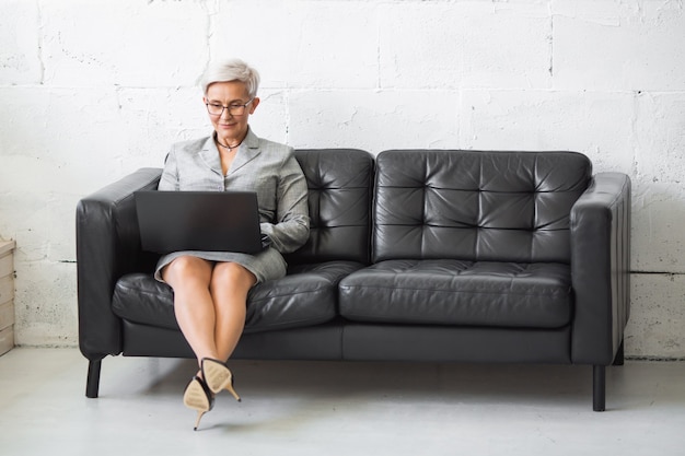 Foto linda mulher adulta elegante em terno trabalhando com laptop no sofá preto