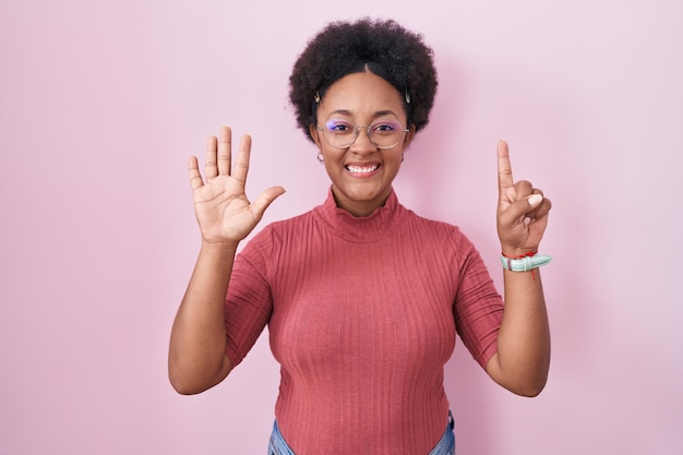 Linda mulher africana com cabelo encaracolado em pé sobre fundo rosa mostrando e apontando para cima com os dedos número seis enquanto sorria confiante e feliz.