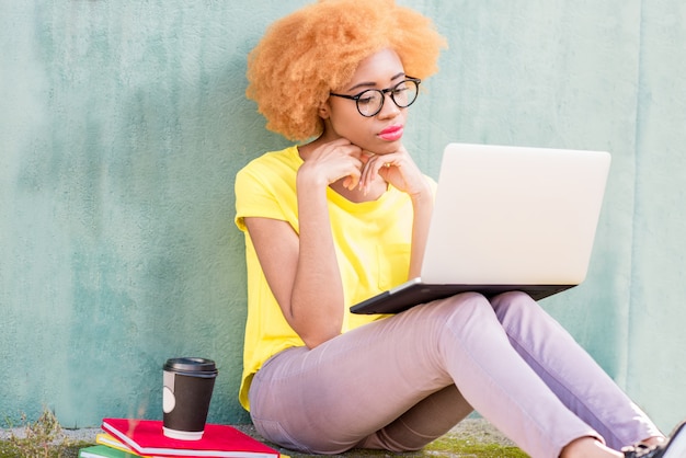 Linda mulher africana com cabelo encaracolado, estudando com laptop e livros sentados ao ar livre no fundo da parede verde.