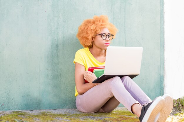 Linda mulher africana com cabelo encaracolado, estudando com laptop e livros sentados ao ar livre no fundo da parede verde.