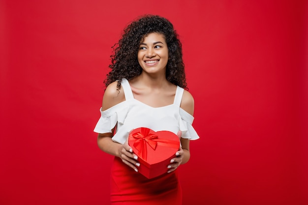 Linda mulher africana feliz segurando o coração em forma de caixa de presente dos namorados isolada em vermelho