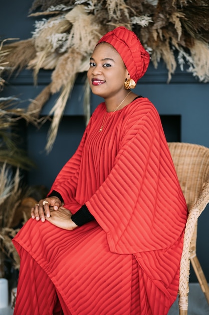Linda mulher africana vestindo um terno vermelho da moda e um lenço na cabeça, sentada na cadeira no estúdio e olhando para a câmera com um sorriso