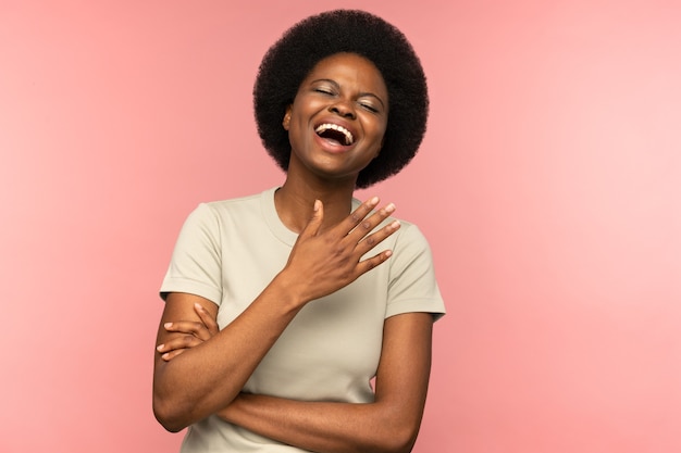 Linda mulher afro-americana com penteado afro rindo de piada com os olhos fechados sobre a parede rosa