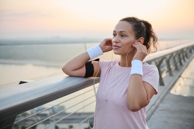 Linda mulher afro-americana esportiva em roupas esportivas, colocando fones de ouvido, ouvindo música, preparando-se para a corrida matinal ao longo da ponte da cidade. Esportista fazendo exercícios ao ar livre ao amanhecer
