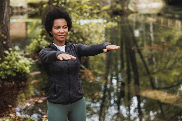 Linda mulher afro-americana madura e sorridente, estendendo-se depois de correr de manhã perto do rio na natureza.