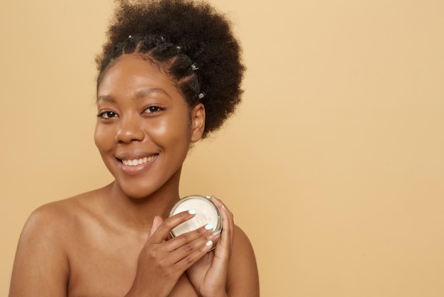 Linda mulher afro-americana segurando o pote de hidratante ou máscara em fundo bege com espaço de cópia Cuidados com a pele antienvelhecimento