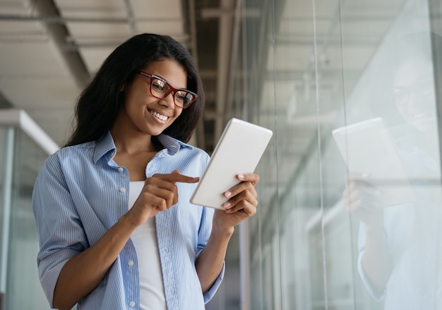 Linda mulher afro-americana sorridente usando tablet digital e trabalhando online em um escritório moderno