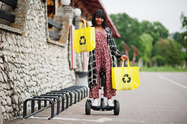 Linda mulher afro-americana usando segway ou hoverboard garota negra com símbolo de reciclagem de sacos ecológicos de pano amarelo