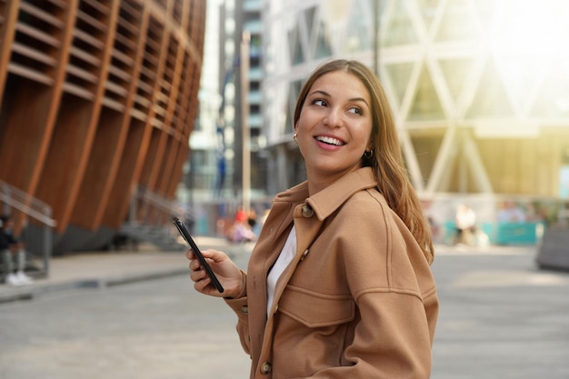 Linda mulher andando na cidade moderna usando o celular olhando de lado na hora do pôr do sol